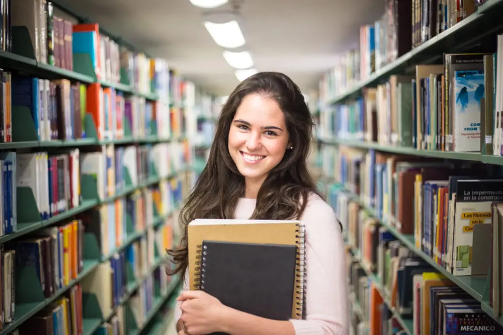 A smiling young woman stands in a library, holding a book, surrounded by shelves filled with various titles