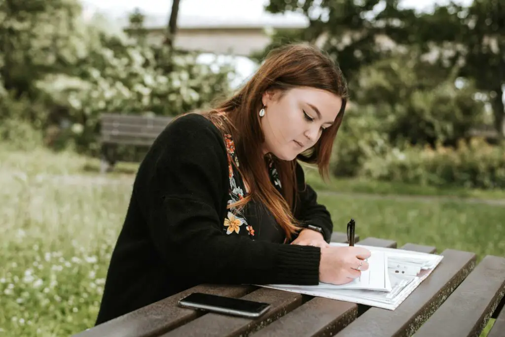 A college student engrossed in writing in her notebook while sitting on a bench to study for college midterm season