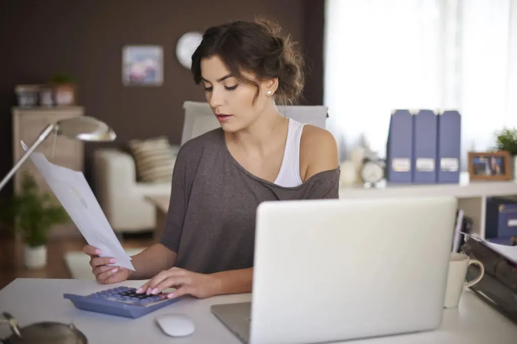 A woman sitting at a desk, working on a laptop and calculating to minimize college expenses