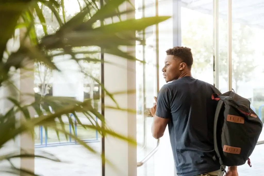 Black student looking outside window carrying black and brown backpack while holding his hand on window