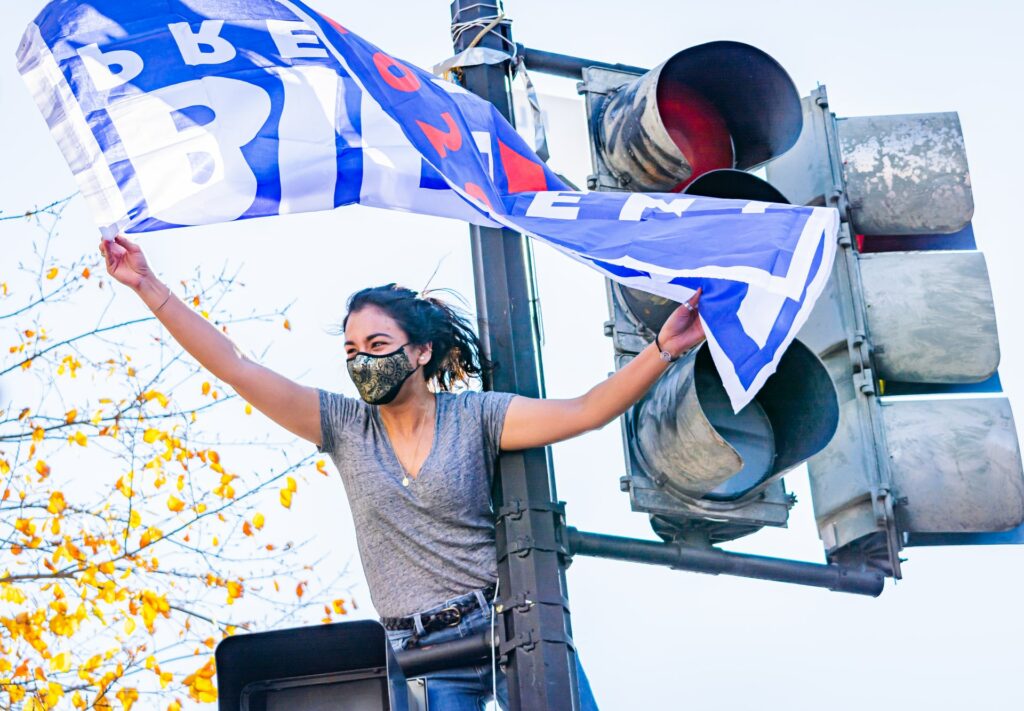 A woman perched on a streetlight holding a Biden-Harris flag