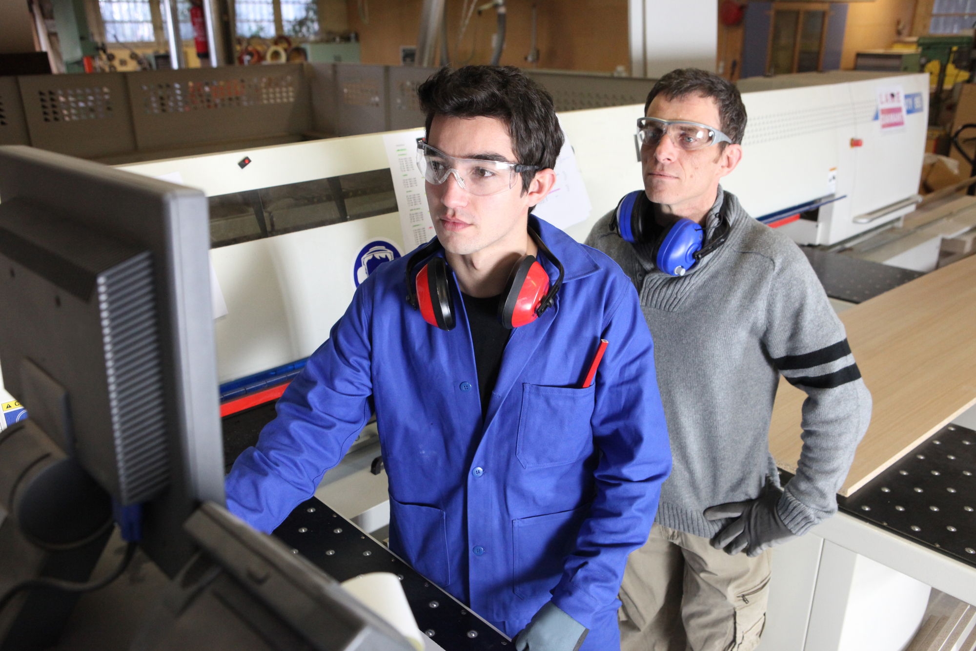 a male professional looks over a male college student wearing safety goggles while working as an intern