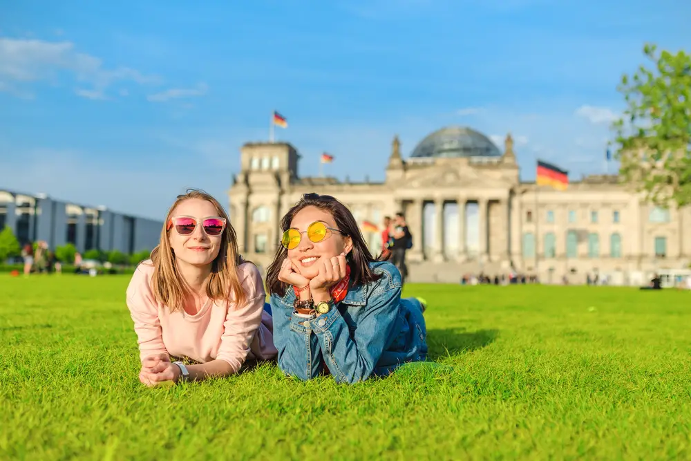 Two young happy female college interns wearing sun glasses lying on the grass and having fun in front of the Bundestag building in Berlin