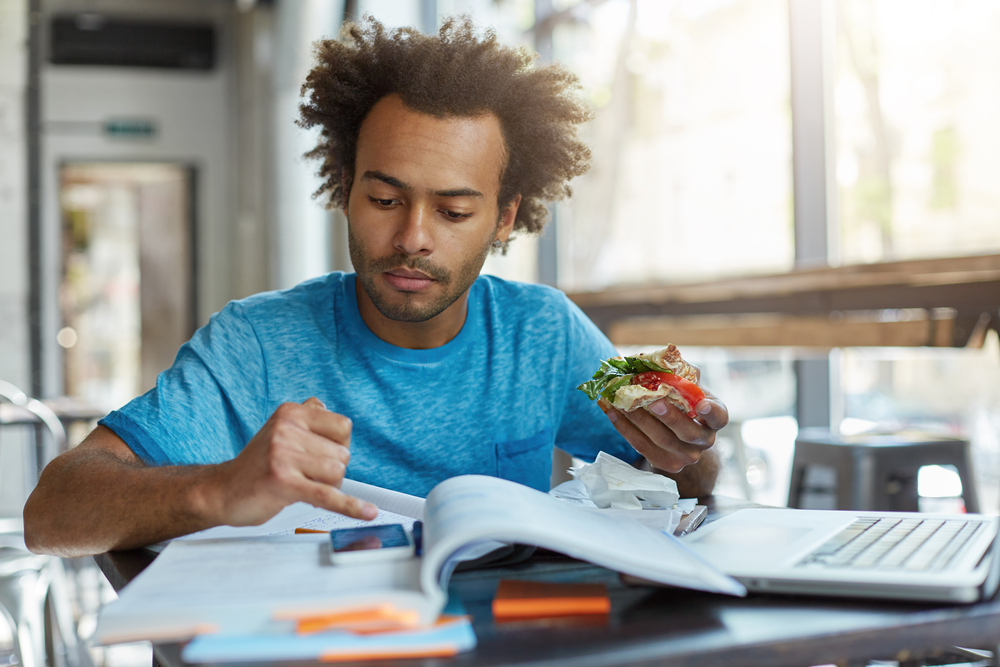 A bushy-haired black student is resting after preparing for class, looking at his smartphone while eating a sandwich