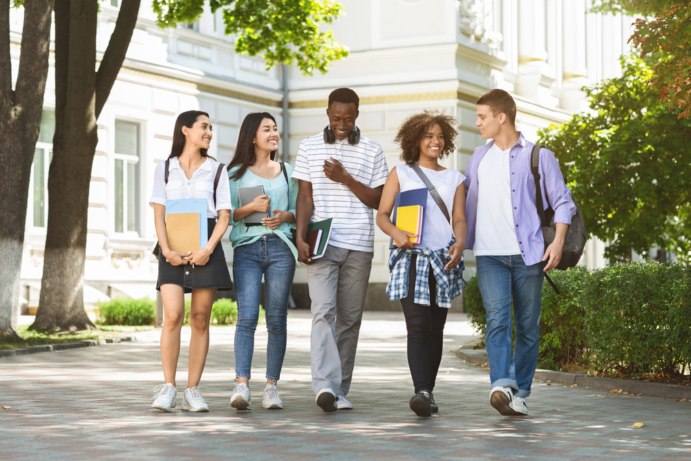 Group of multiethnic students walking together outdoors in college campus, holding books and notepads and laughing