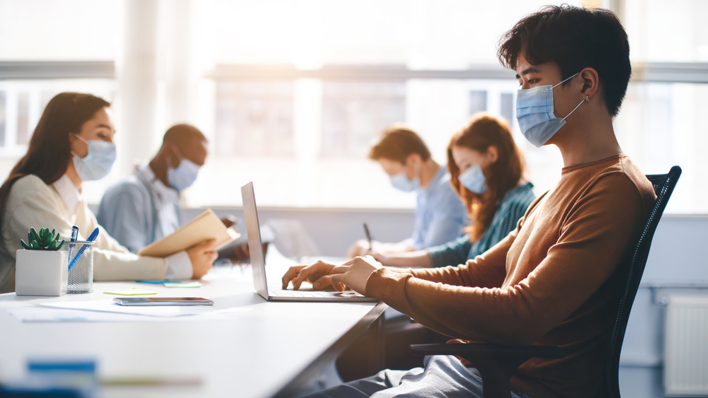 Group of diverse students wearing protective medical masks and using laptops to study