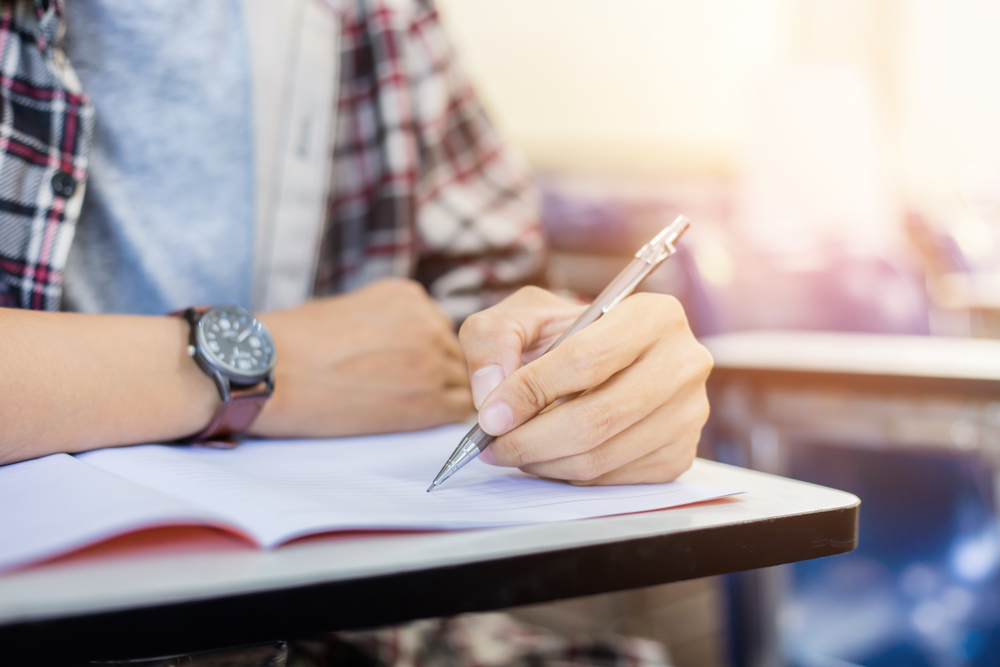 University student holding pencil while writing on exam answer sheet in college hall