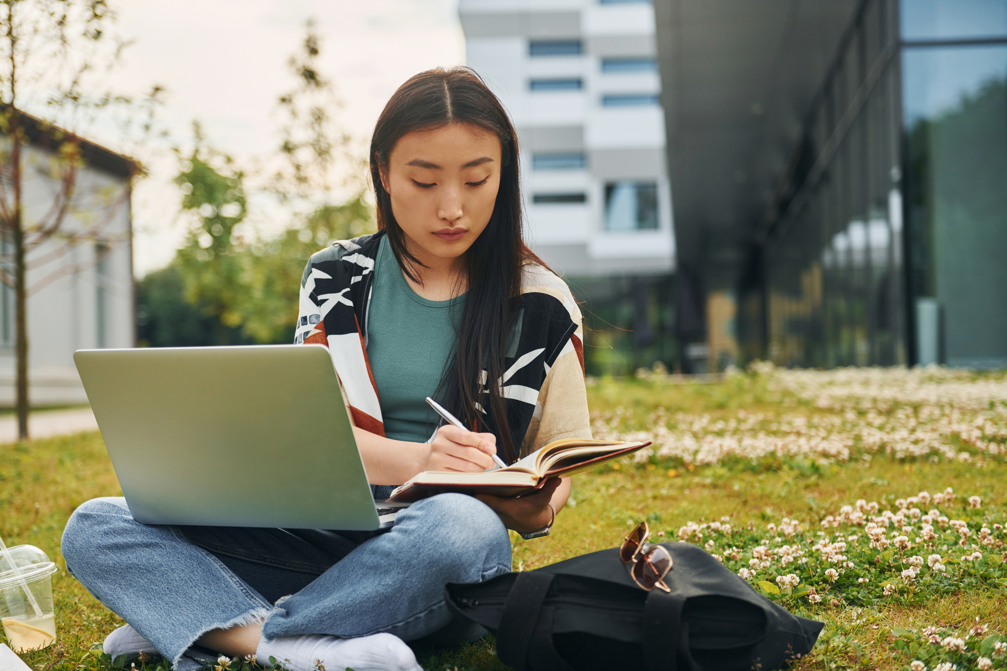 A female college student sitting outside studying with a book and laptop