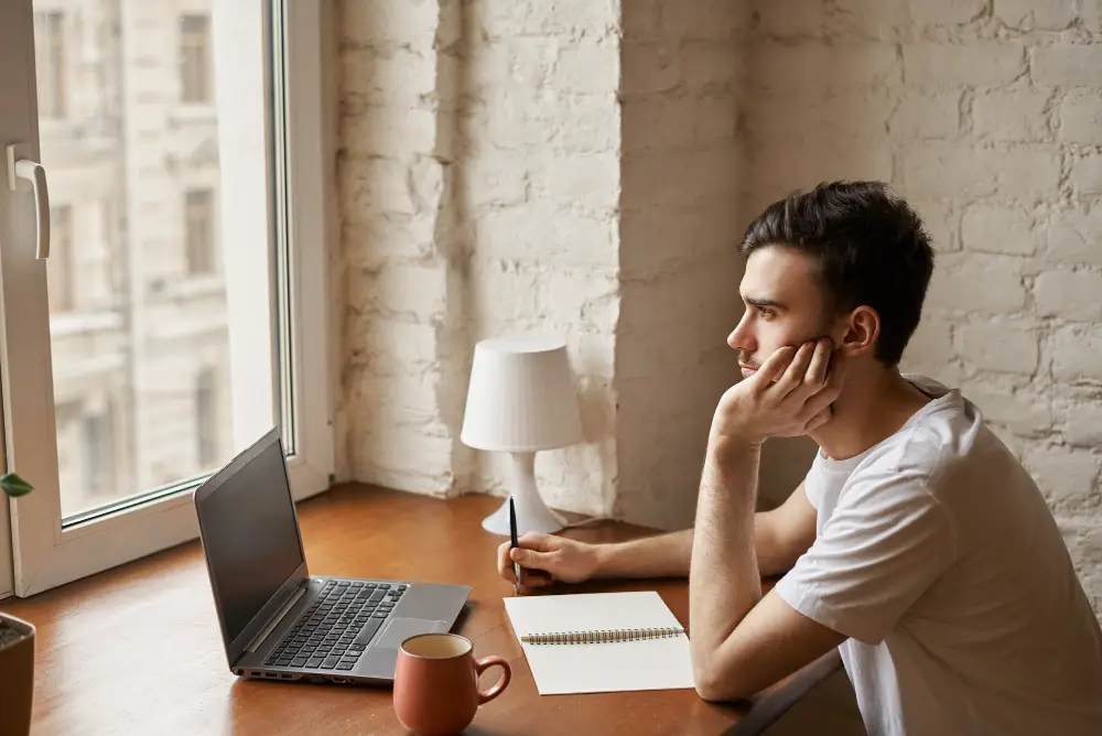 A young man thinks about whether to attend tuition free colleges or go the alternative route in front of his laptop 