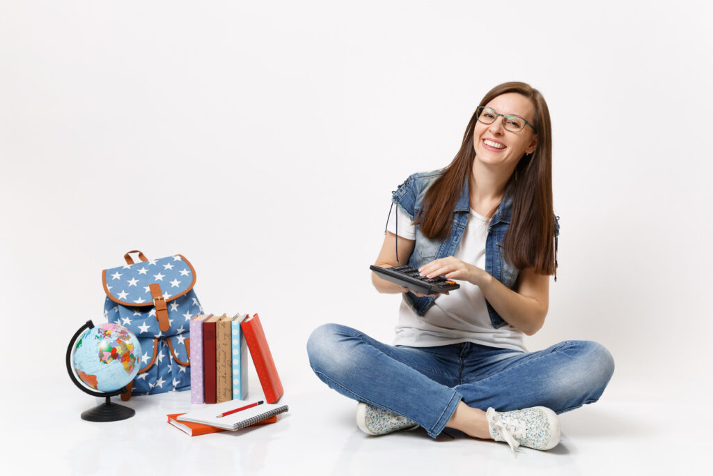 Young laughing woman student holding and using calculator solving math equations sitting near globe, backpack, school books