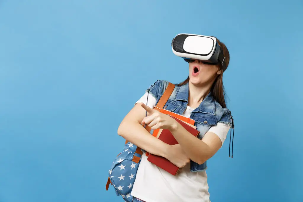 Young shocked woman student with opened mouth with backpack in virtual reality glasses hold school books pointing index finger isolated on blue background