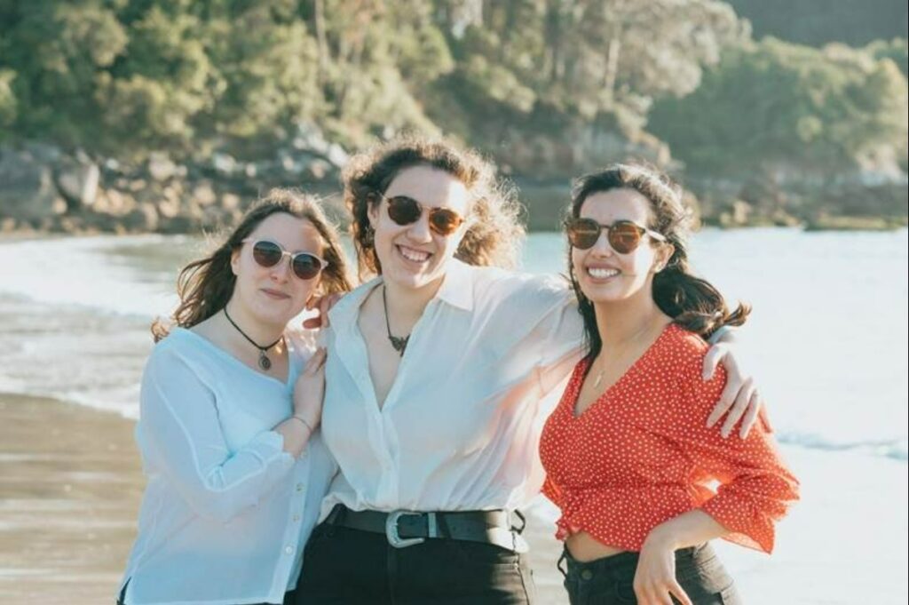 Three women smile at the camera on the beach.