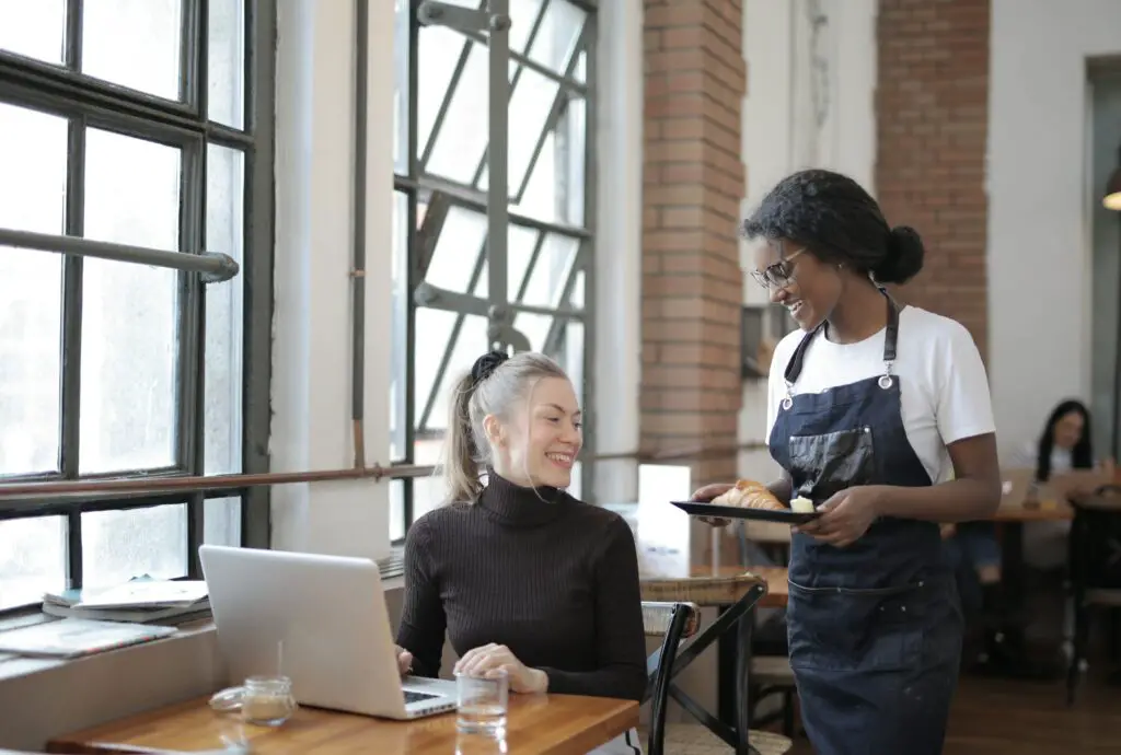 african-african-server-speaking-to-guest-who-is-working-on-her-laptop-at-restaurant