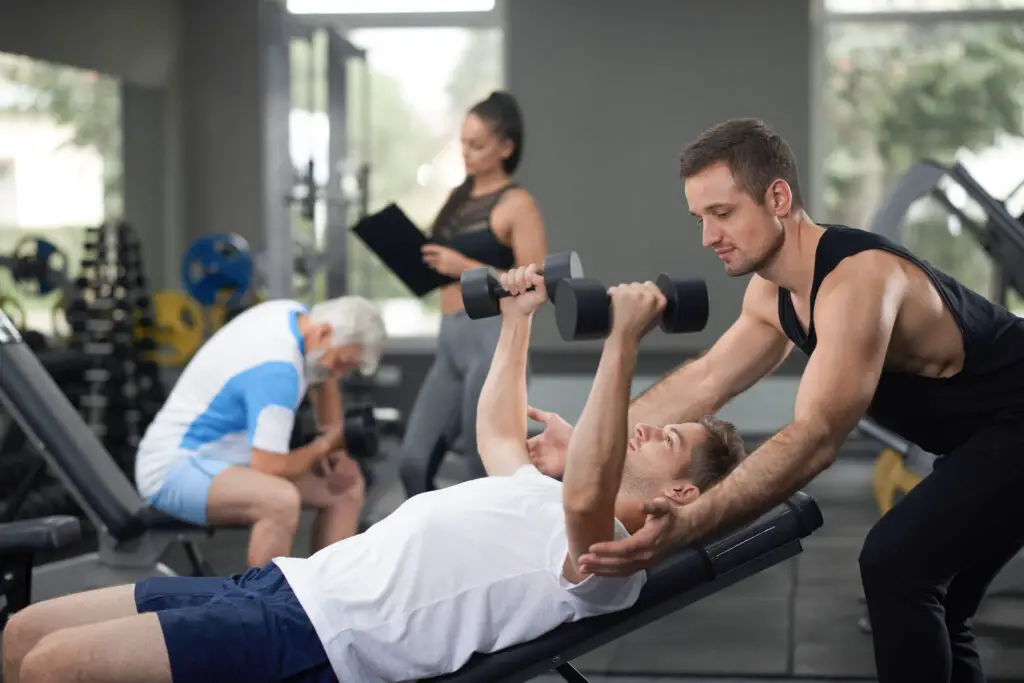 College male part-time gym attendant assisting gym goer lifting dumbbells