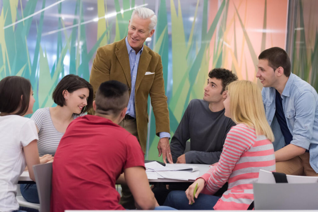 Male professor smiling while discussing the alternative grading system using contracts with his six students around a round table