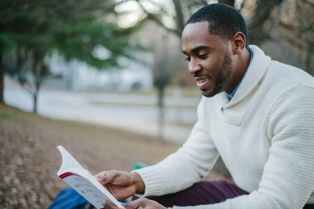 African-American college student in white sweater reading a book outside with trees in background