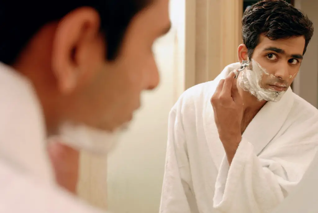male-college-student-in-a-bathrobe-using-a-razor-from-his-college-packing-checklist-to-shave-his-beard-in-front-of-a-mirror