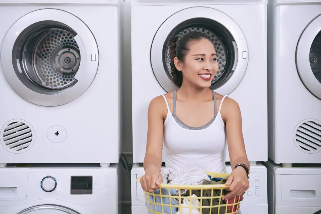 female-college-student-in-front-of-washer-machines-holding-a-laundry-basket-with-clothes-from-her-college-packing-checklist