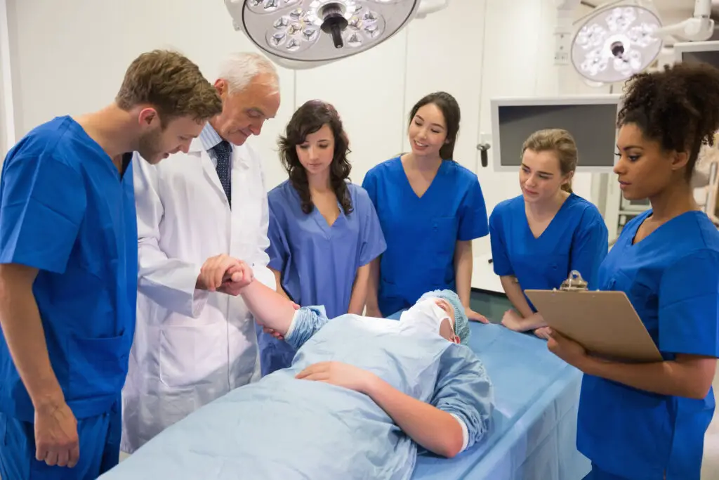 male and female medical students in blue scrubs with a doctor wearing a lab coat stand around a patient and discuss symptoms using the prognosis study tool