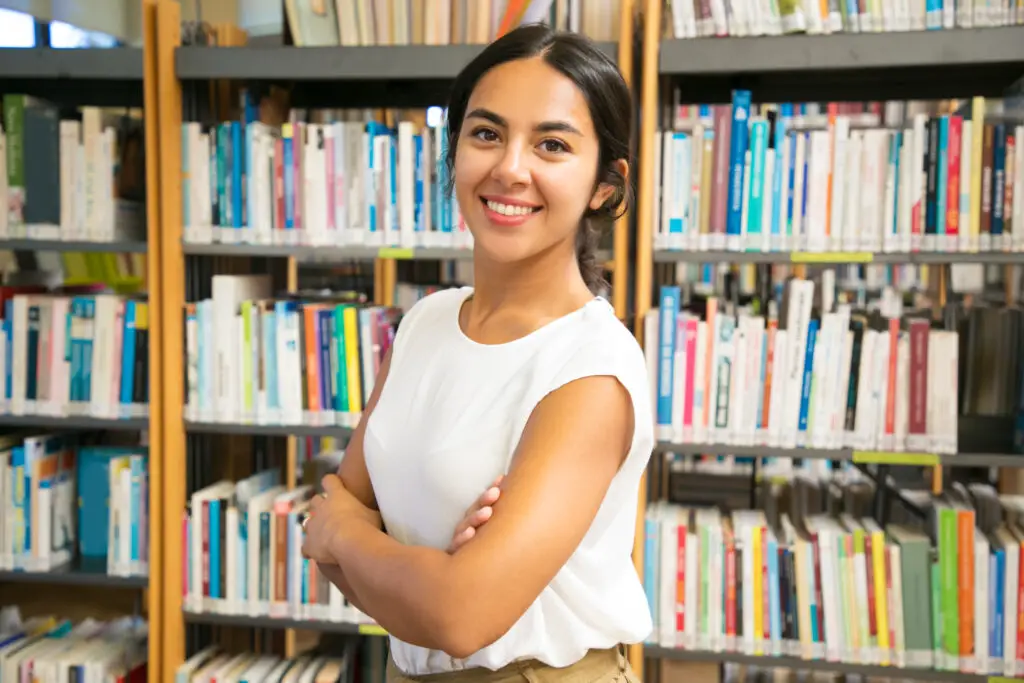 A smiling Latina law student from the Universidad de Morón, among the weirdest college names worldwide, poses with her arms crossed in the campus library