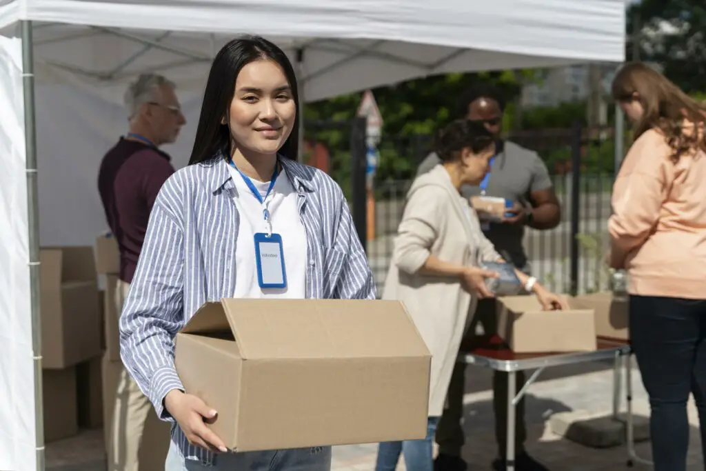A smiling Asian female college student carries a box filled with packed meals for a food bank volunteering organization 