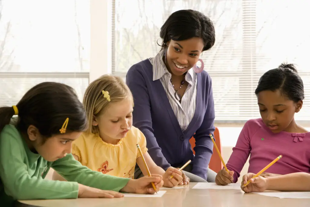 a-female-teacher-wearing-a-purple-cardigan-sits-with-her-class-of-young-girls-and-engages-them-in-an-activity-using-pencils-and-papers