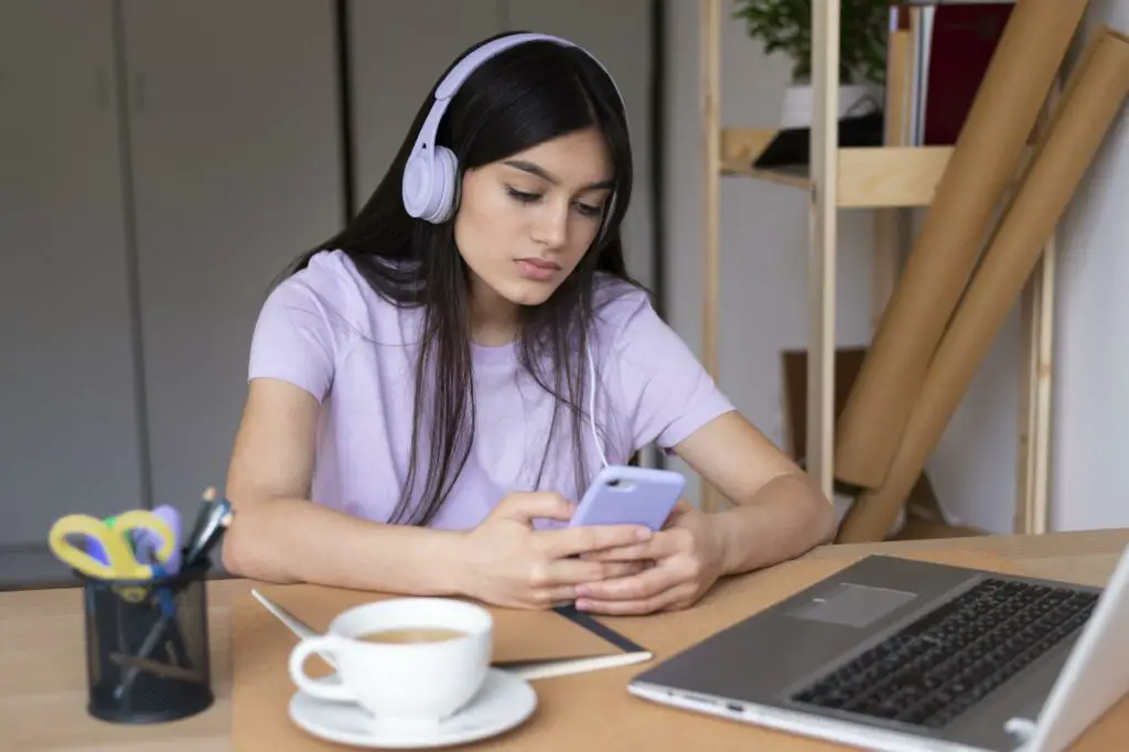 A female college student wearing headphones is distracted by her phone instead of listening during online class