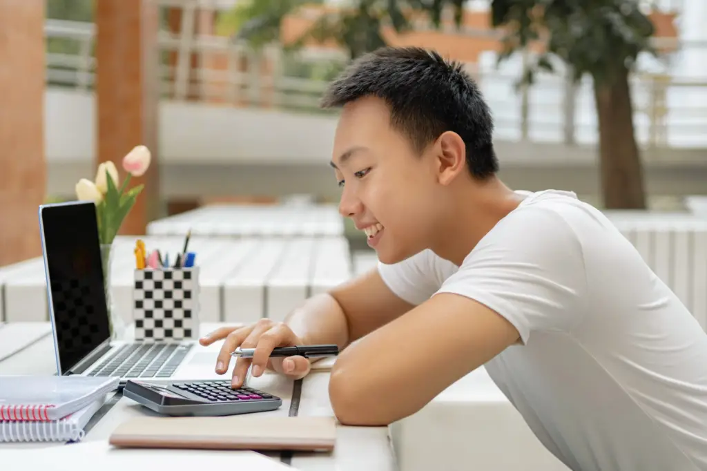male international student using a calculator pen and laptop on a desk to prepare a budget for studying college in the US