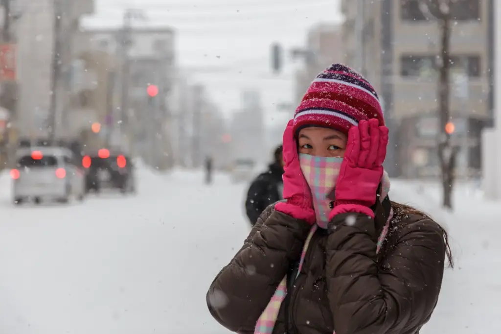a female international college student wearing pink winter gloves and puff jacket covering her ears in the middle of snow-covered street