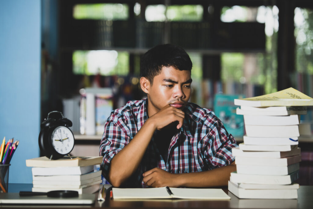 An Asian male high school graduate deciding between attending college vs trade school after reading resources in the school library
