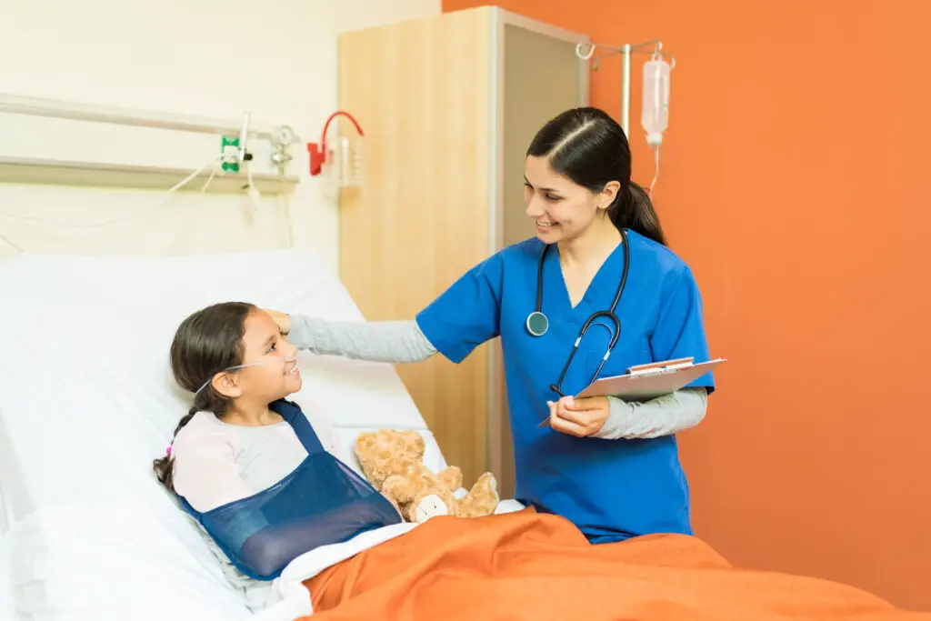 A smiling raven-haired nurse checks the girl patient with an arm sling lying on the hospital bed