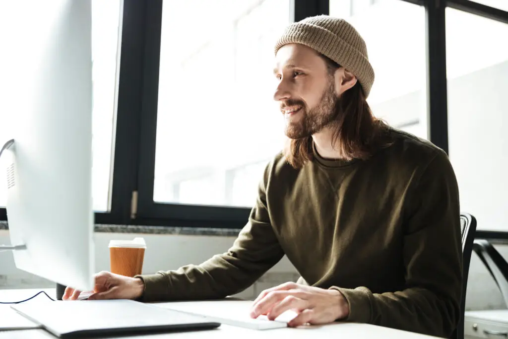 A smiling long-haired male web designer wearing a beanie arranges website elements on the office desktop