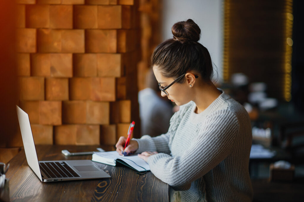 a female college student reading a book and sitting in a library while using online study tools on her laptop