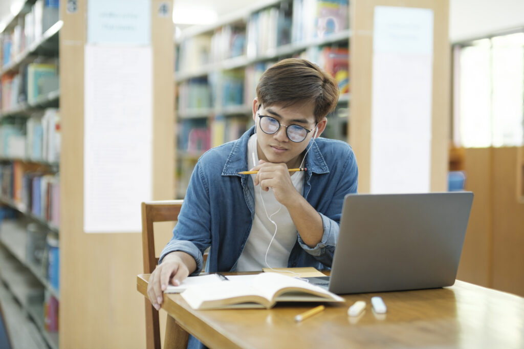 a male college student wearing earphones sitting in a library while using online study tools