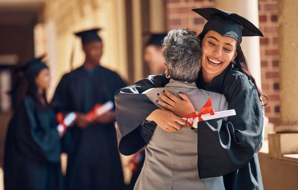 a female college student wearing a graduation toga and holding a diploma hugs her mother while smiling