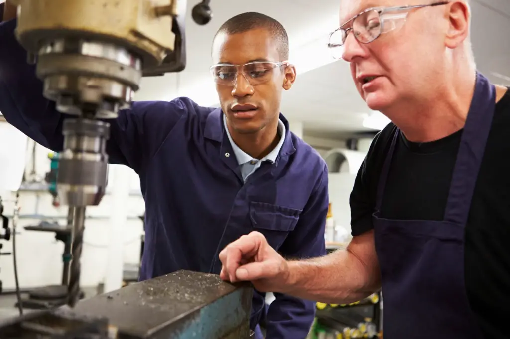 a male college student in an apprentice program and his male mentor wearing safety goggles in front of a machine