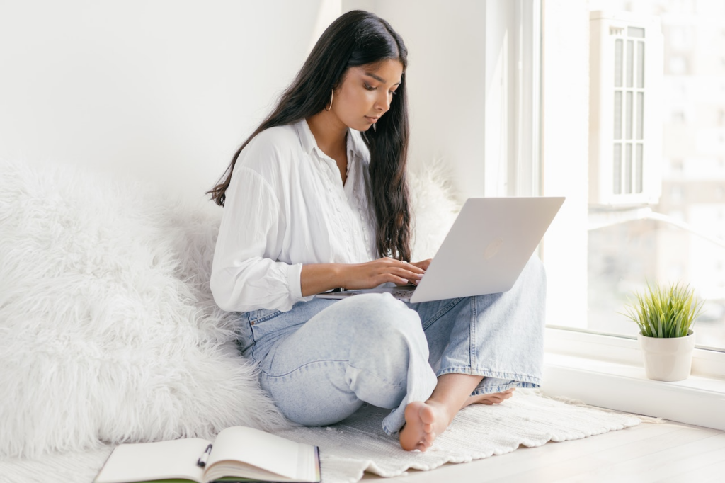 a female college student sits by the window at home while studying on her laptop