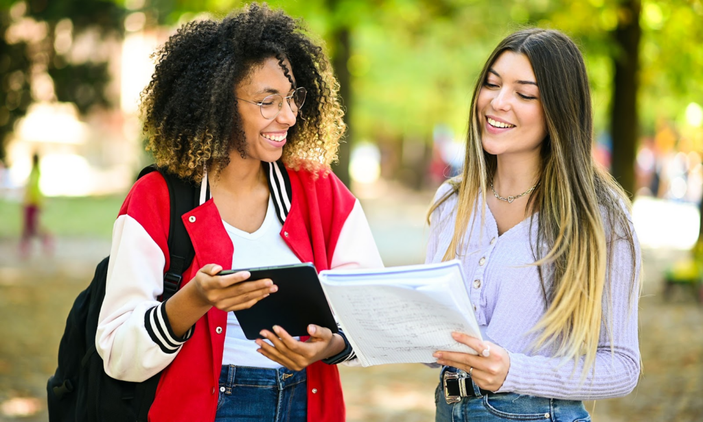 two female high school students enrolled in summer class are comparing their notes outdoors