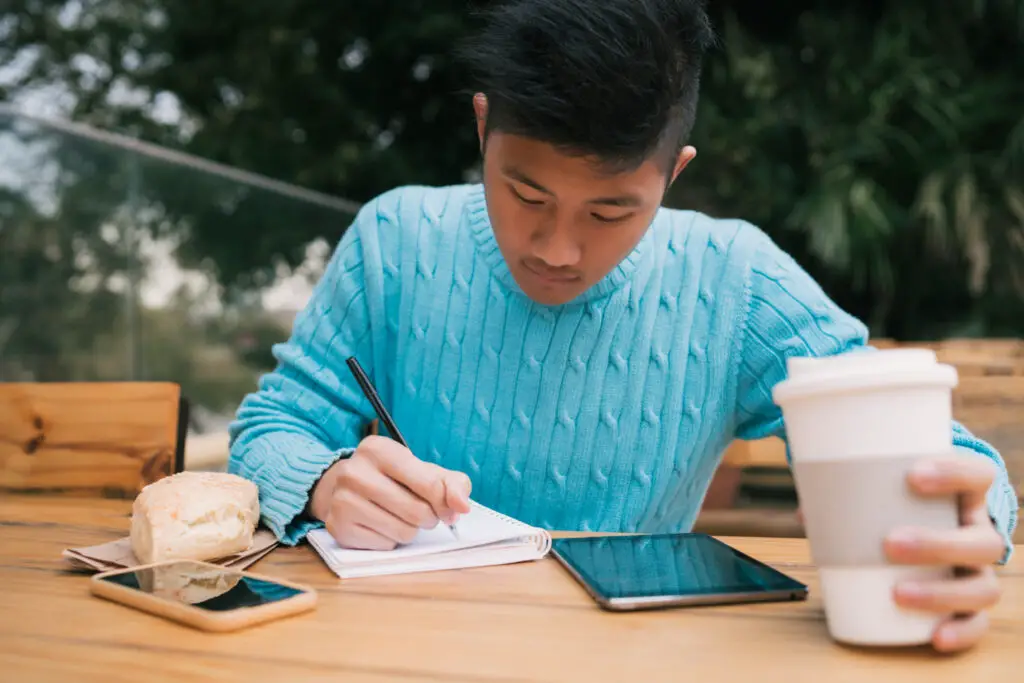 asian male college student wearing blue sweater holding coffee cup writing on his notebook