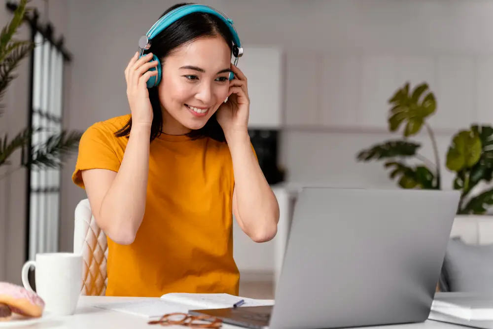 A smiling Asian female student wearing headphones is attending an online class on her budget laptop
