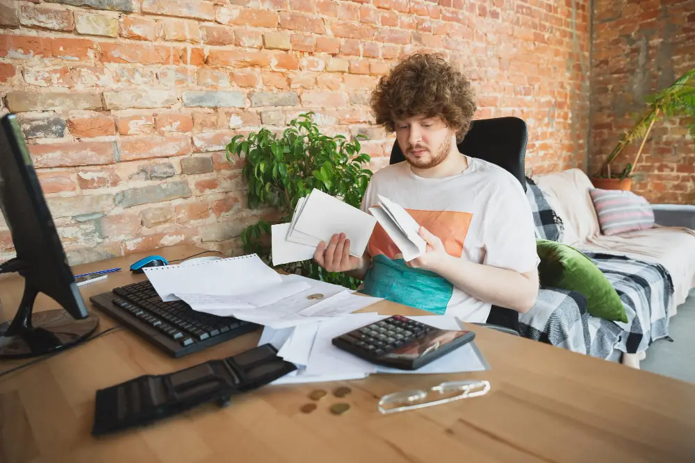 A Caucasian curly-haired male college student holds his bills and cash envelopes as he figures out how to budget in college