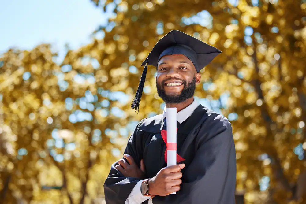 male college graduate wearing a diploma cap and gown proudly holding his diploma