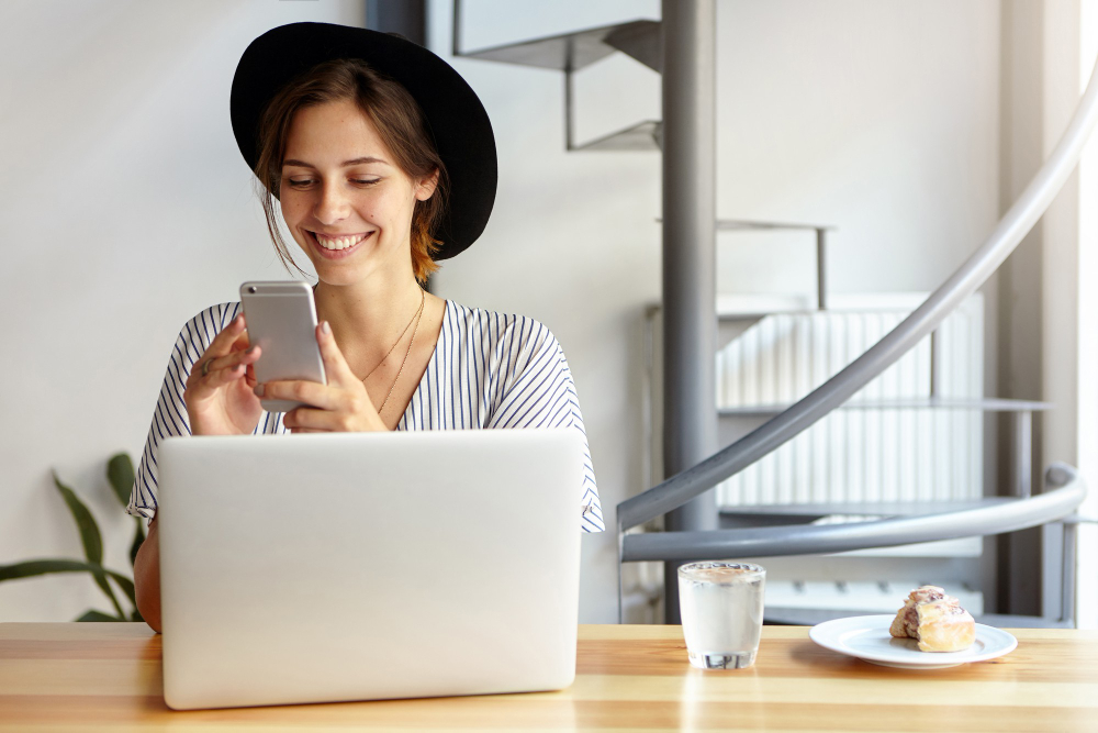 A smiling female social media manager checks a company's online platform on her phone and laptop
