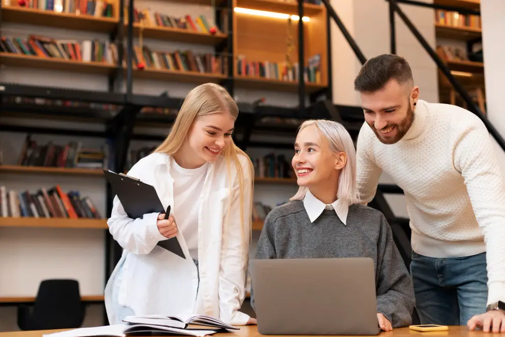 A group of three smiling blonde college students are studying and comparing notes in the library