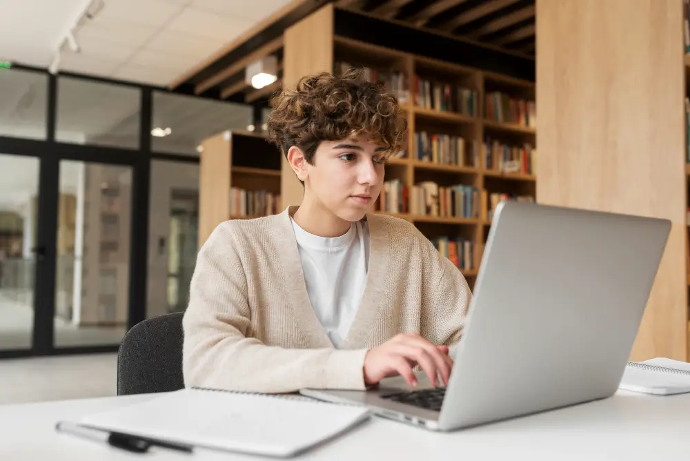 A short curly-haired college student focusing on research on their laptop in the library as part of their preparations for a law school routine