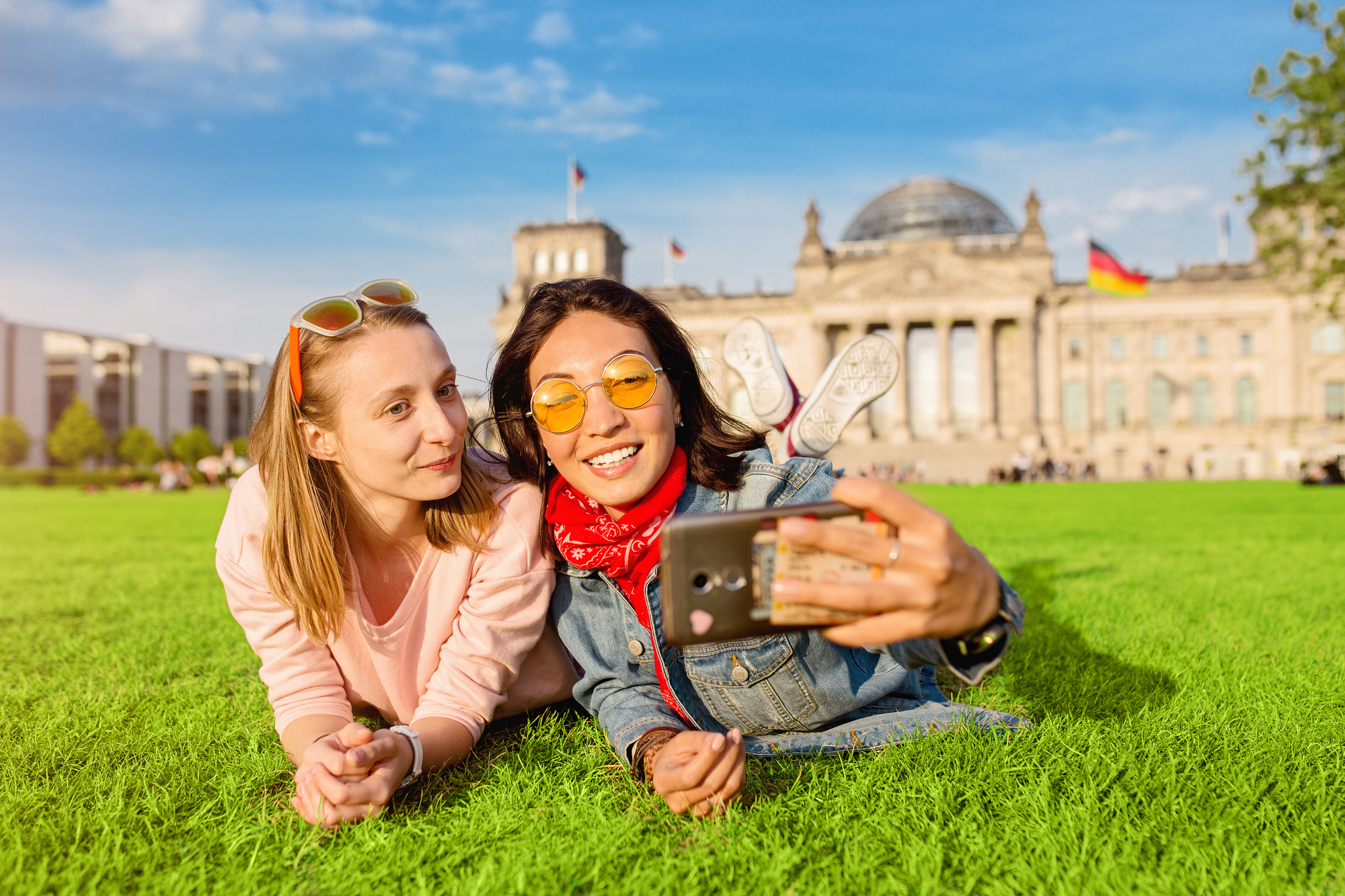 two-happy-young-girls-lying-on-grass-take-selfie-with-historic-building-and-german-flag-in-the-background-during-study-abroad-program