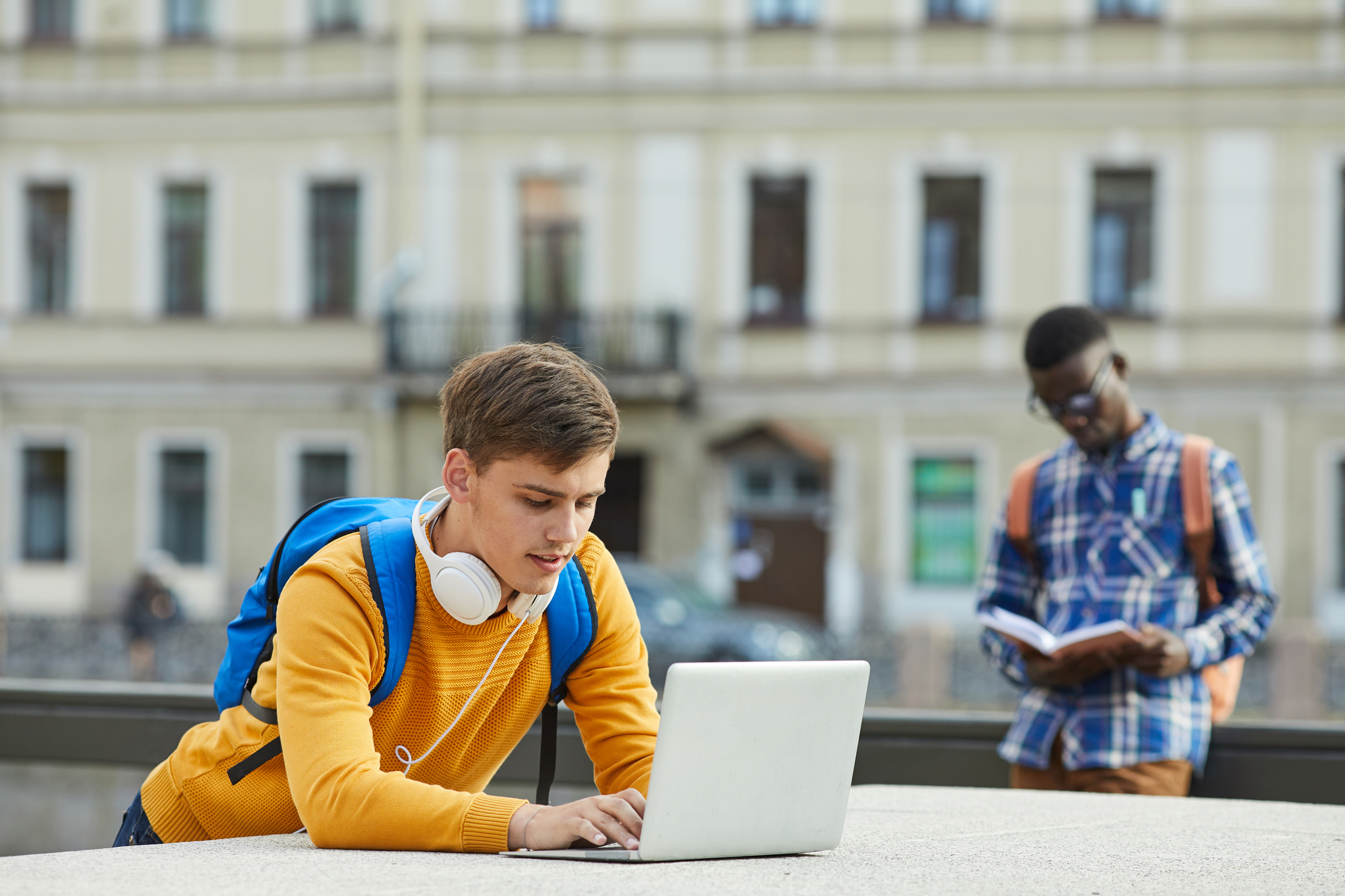 male-college-students-working-on-a-laptop-and-reading-a-book-outdoors-during-overseas-study