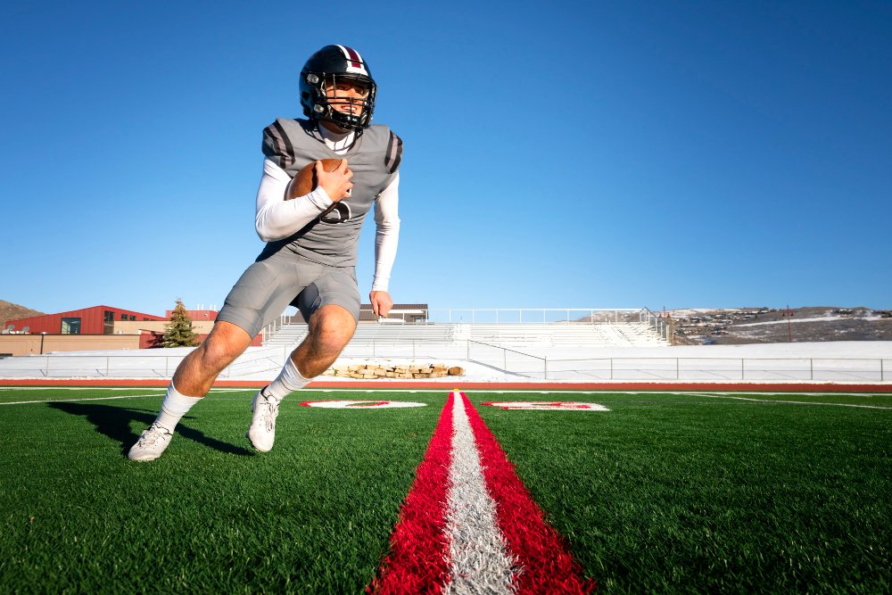 A male American college footballer runs while holding a ball on the green outdoor field