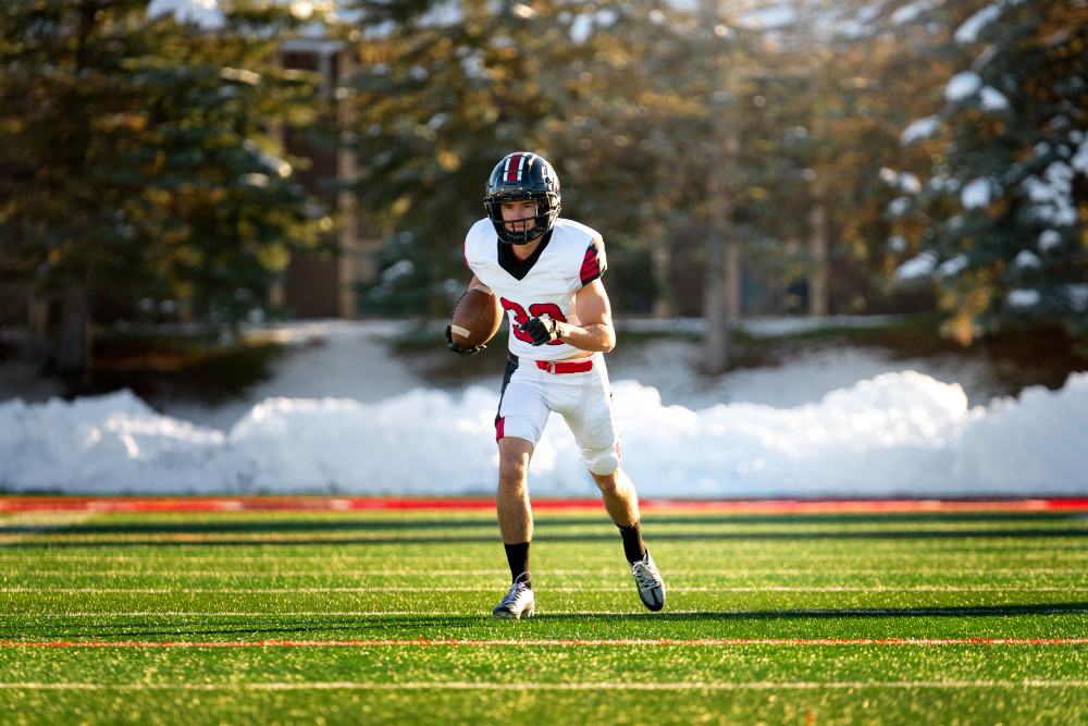 A male American football player runs across the field carrying the ball