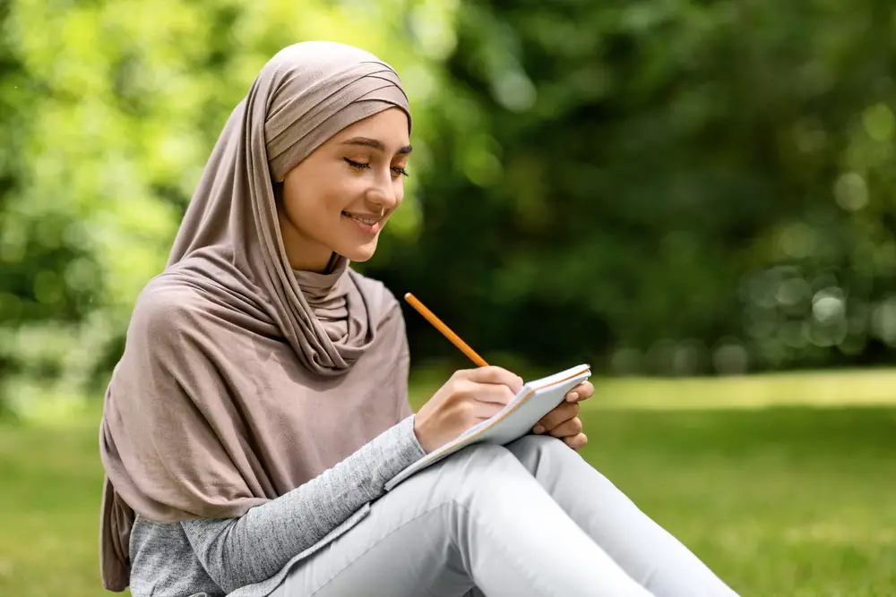 female college student wearing hijab writing her college essay outdoors